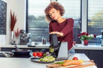 Woman using a kitchen device to cut a cucumber with other vegetables and a pan on top of the countertop